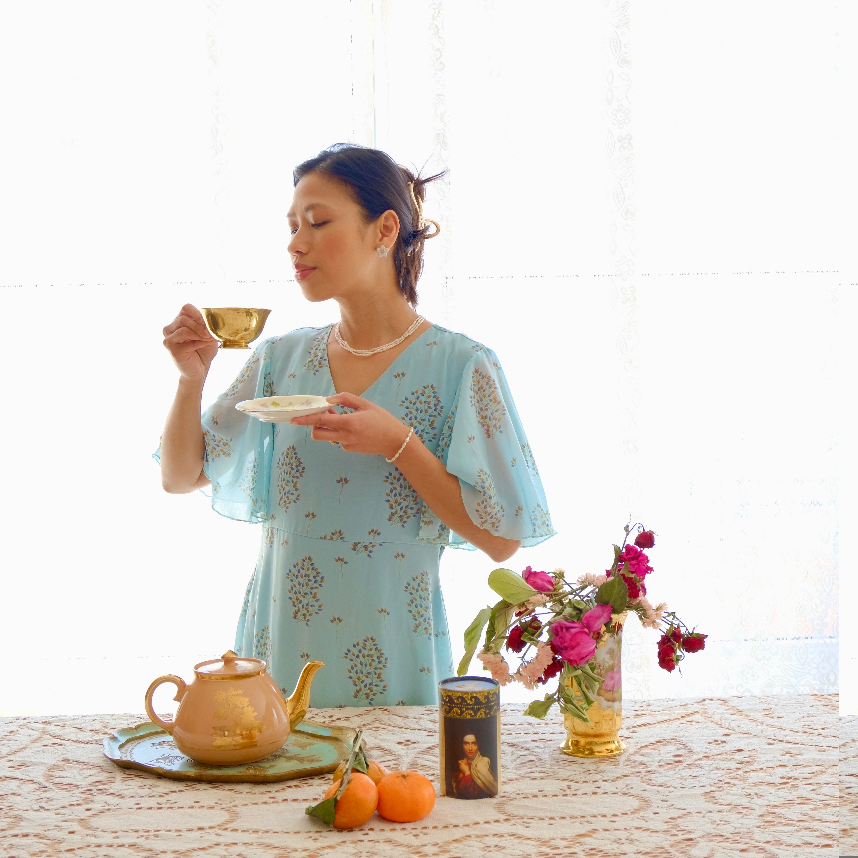 Woman in a light blue dress holding a teacup with a tea set on a table.