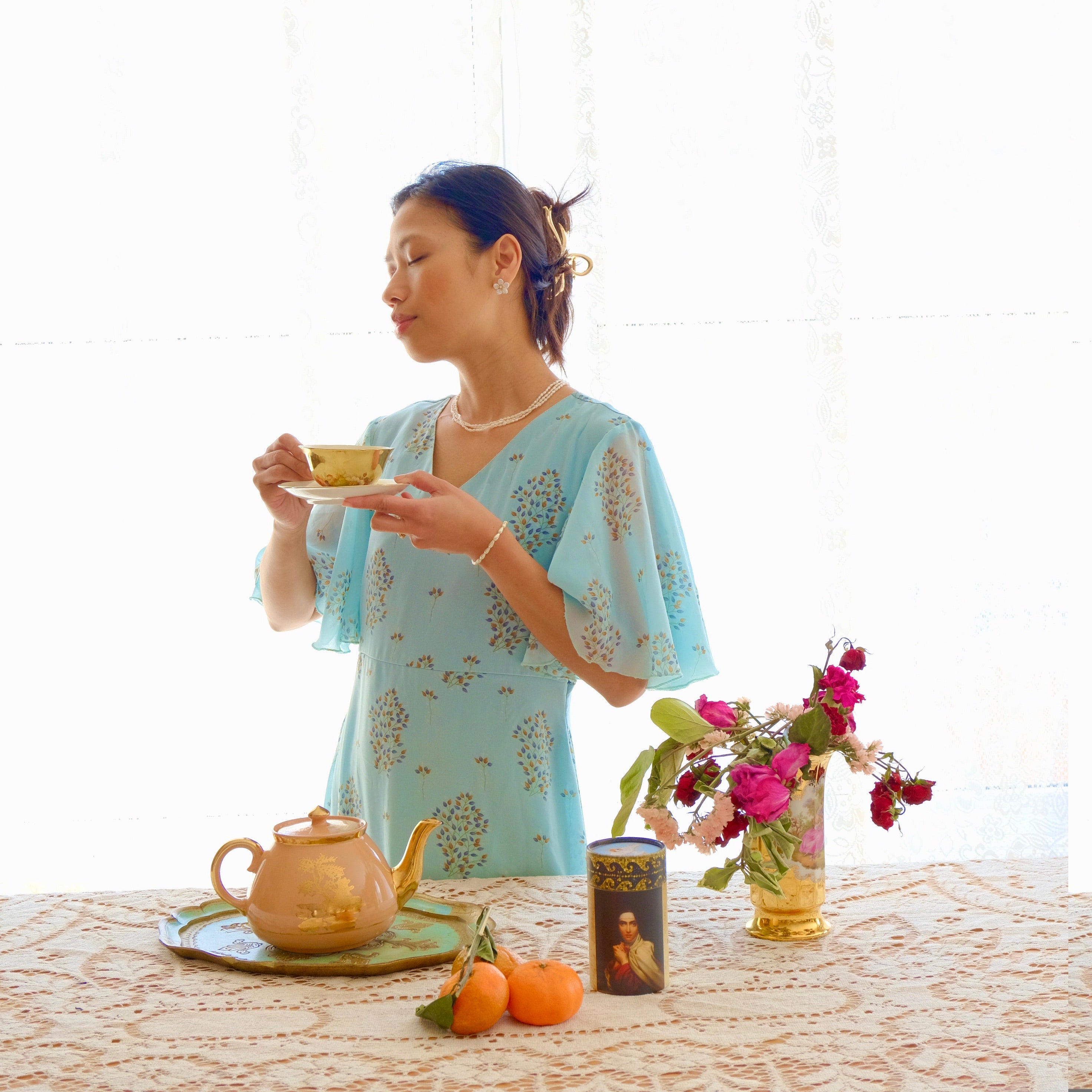 Woman in a light blue dress holding a teacup with a tea set and flowers on a table.