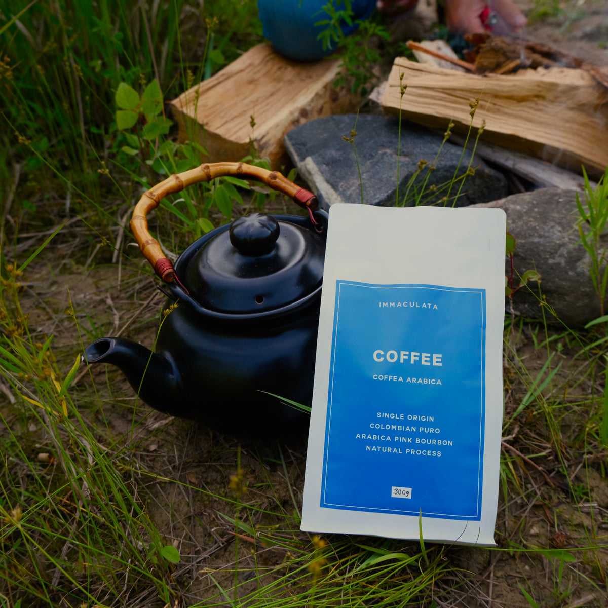 Black teapot and coffee package on grass with firewood in the background