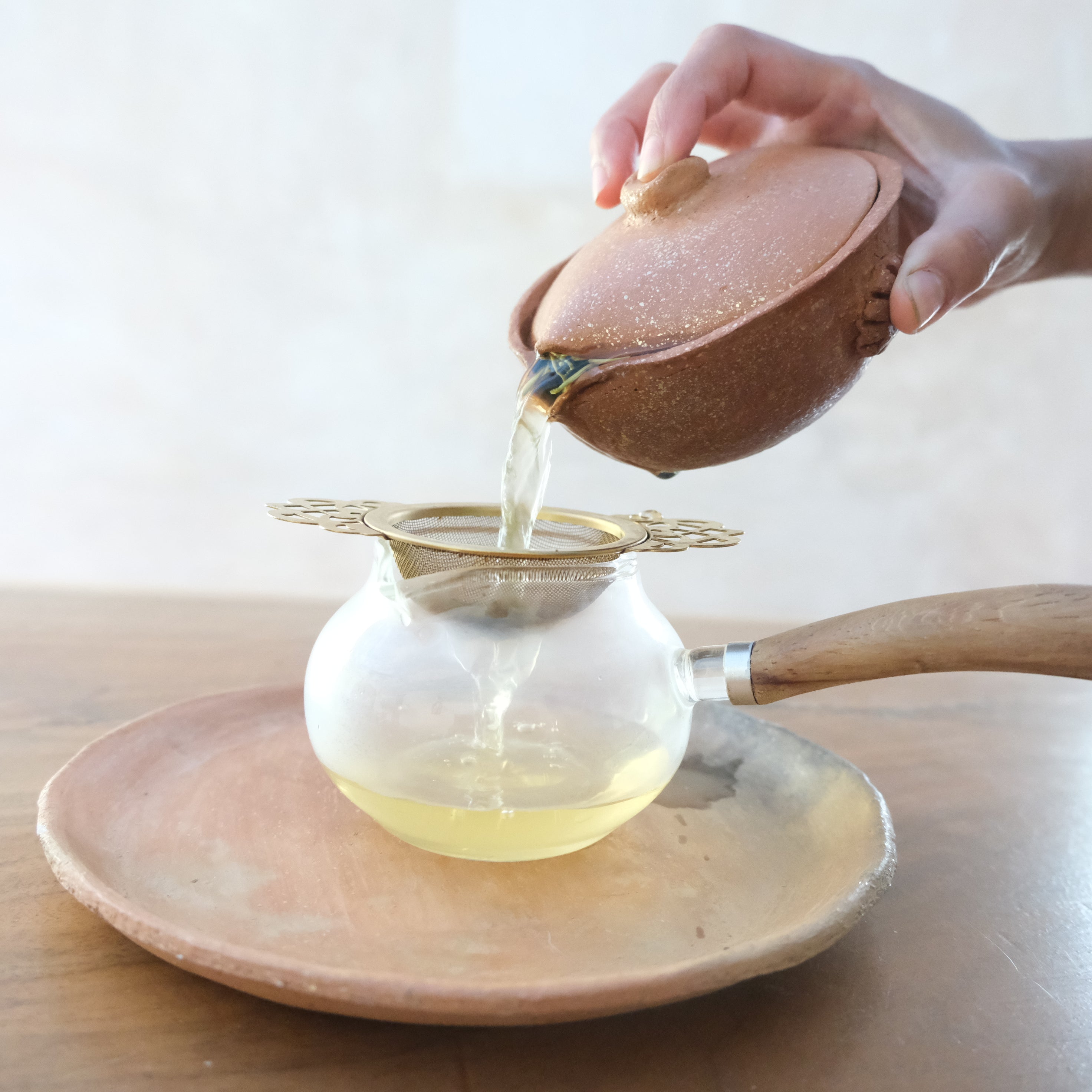 Person pouring tea from a ceramic teapot into a glass teapot with a wooden handle on a light background.
