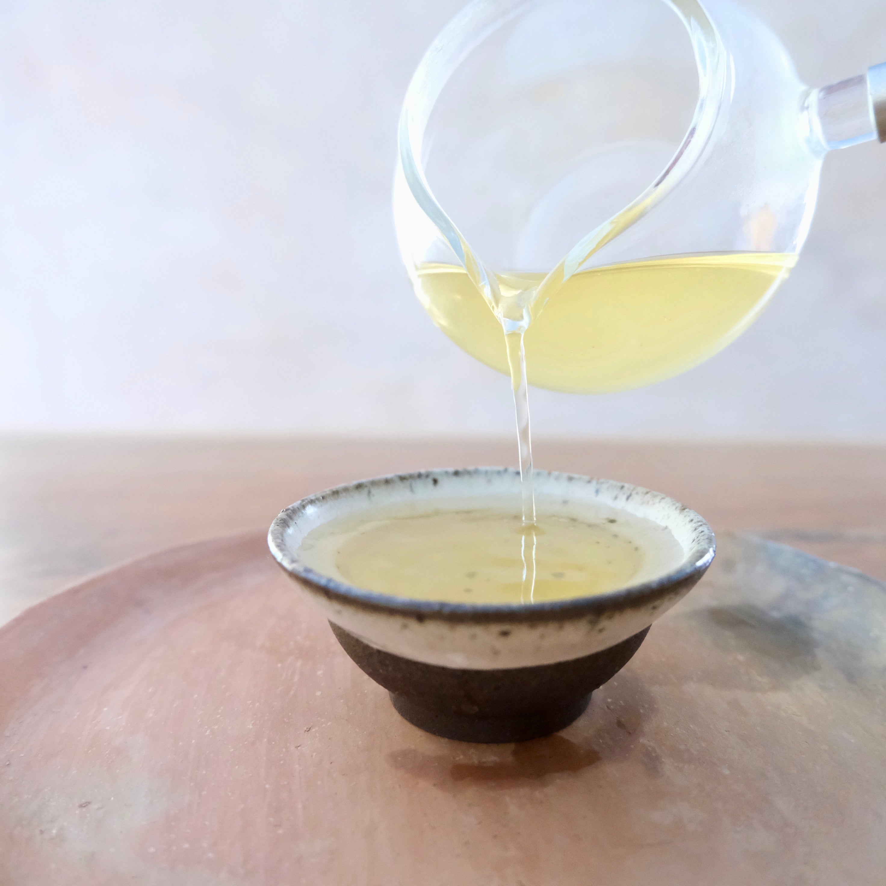 Kukicha Tea being poured from a glass teapot into a ceramic cup on a neutral background