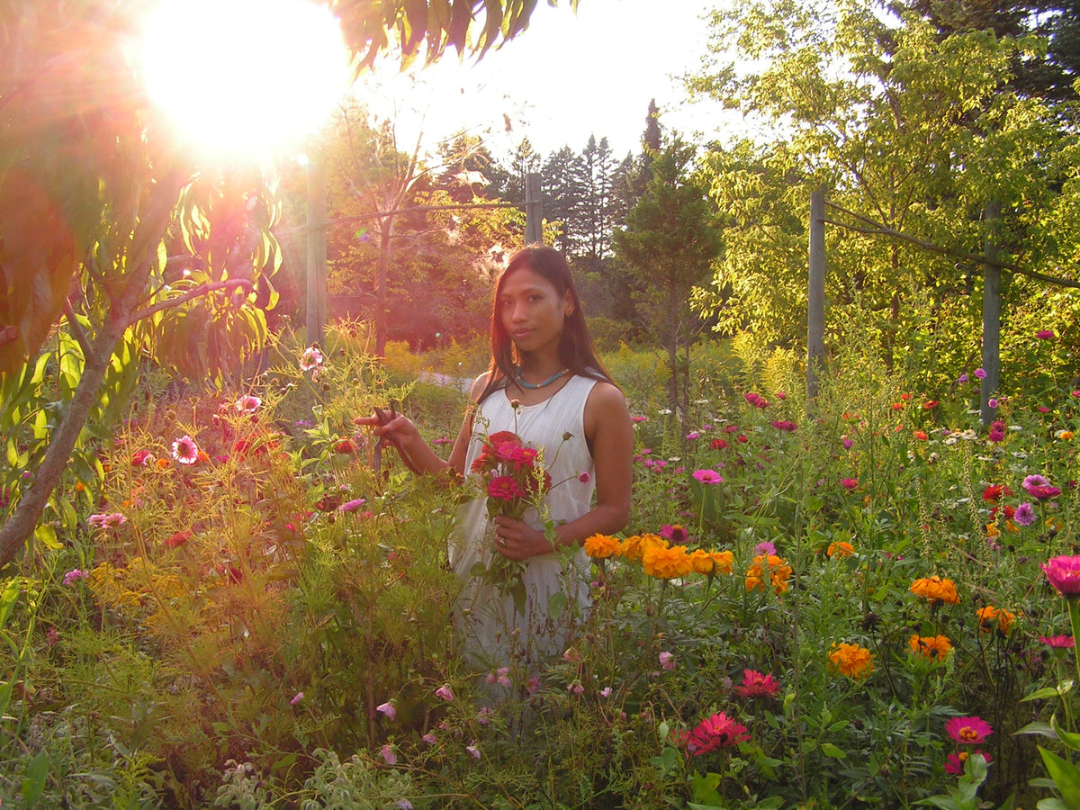 A woman stands in a sunlit flower garden holding a small bouquet of pink flowers. She is wearing a sleeveless white dress, surrounded by green foliage and colorful flowers including orange and pink blooms.