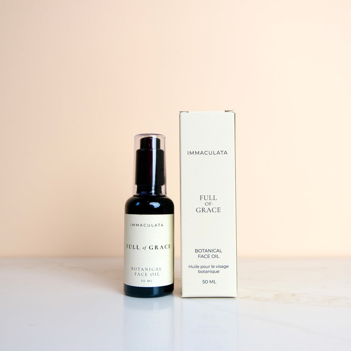 Immaculata Full of Grace Botanical Face Oil 50ml bottle in a dark amber glass bottle with a black pump, standing next to its cream-colored cardboard packaging box on a white surface against a warm beige background.