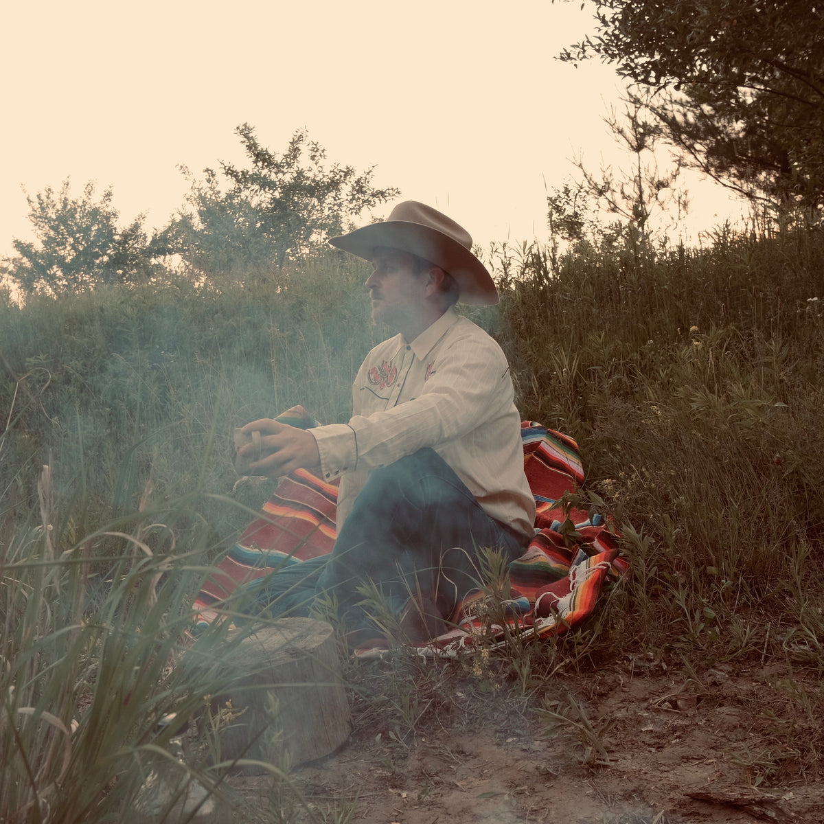 Person sitting on a colorful blanket in a natural setting with trees and grass.