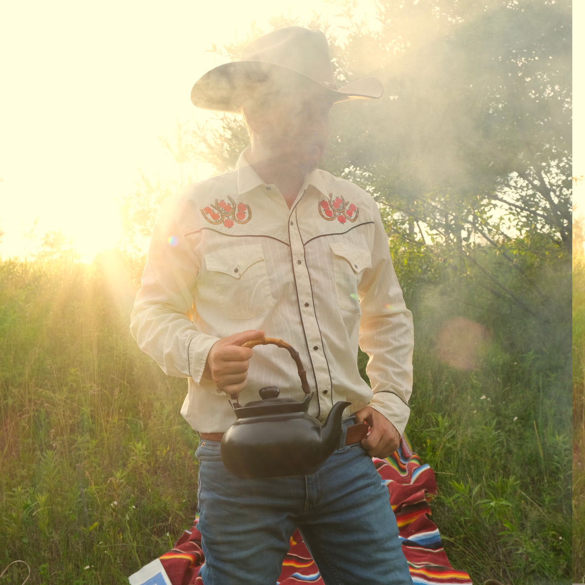 Person in a cowboy hat and embroidered shirt holding a teapot in a field with a blurred background