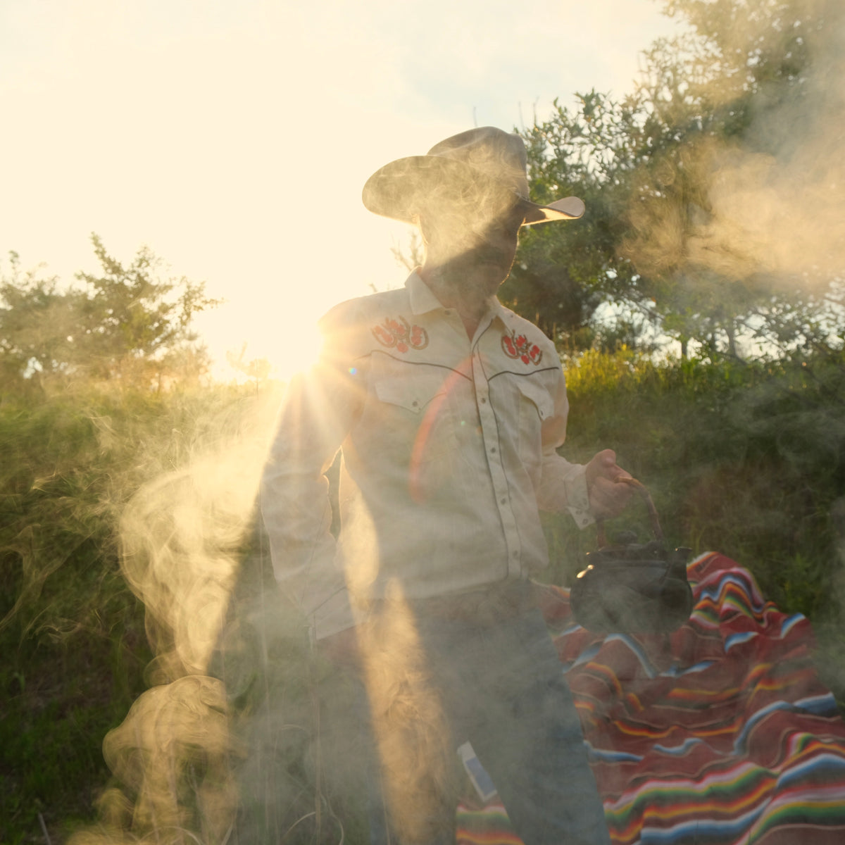Person in a cowboy hat and patterned shirt standing outdoors with smoke and trees in the background