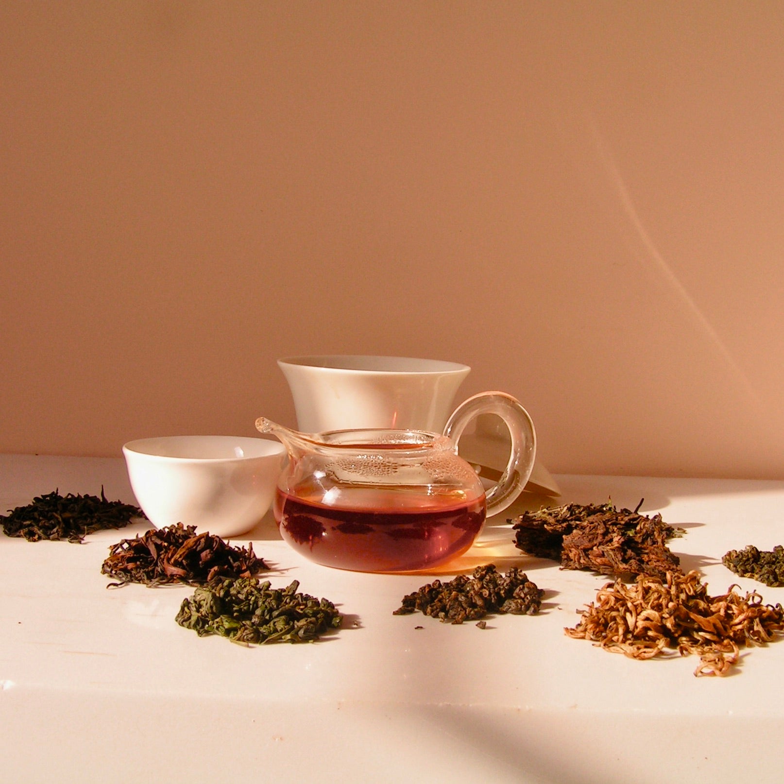 Tea set with a glass teapot filled with tea, surrounded by dry tea leaves on a light surface.