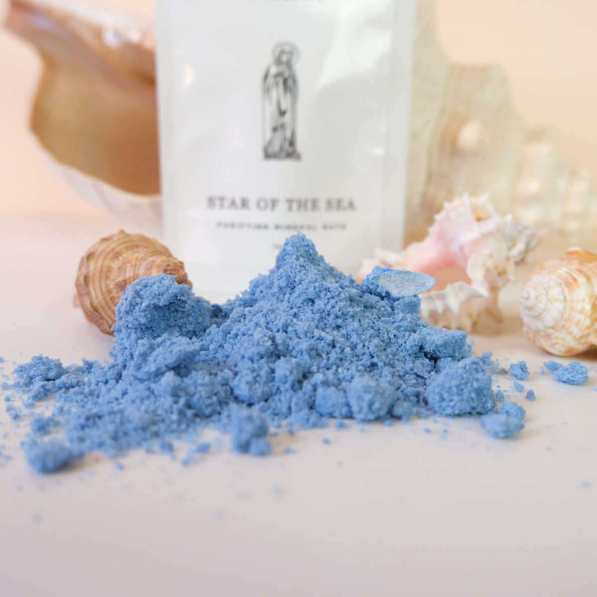A pile of bright blue powder, likely bath salts or mineral powder, displayed in the foreground with a white pouch labeled 'STAR OF THE SEA' and several seashells in the background.