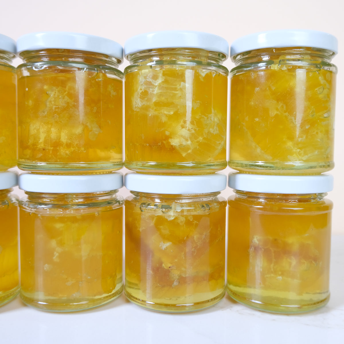 Nine glass jars filled with golden yellow honey or honeycomb pieces, arranged in two rows on a light background. The jars have white lids.