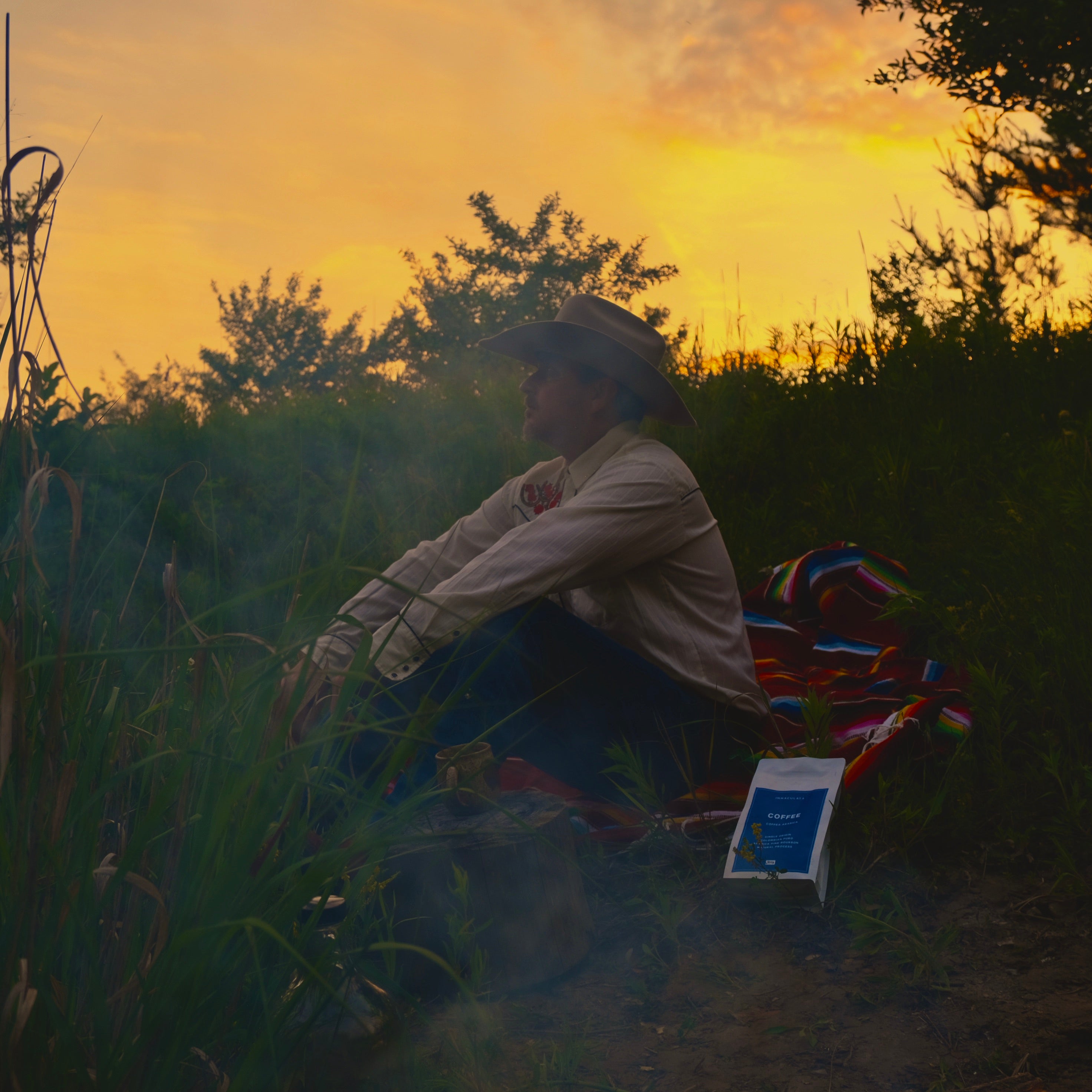 Person sitting in a field with a colorful blanket and coffee at sunrise