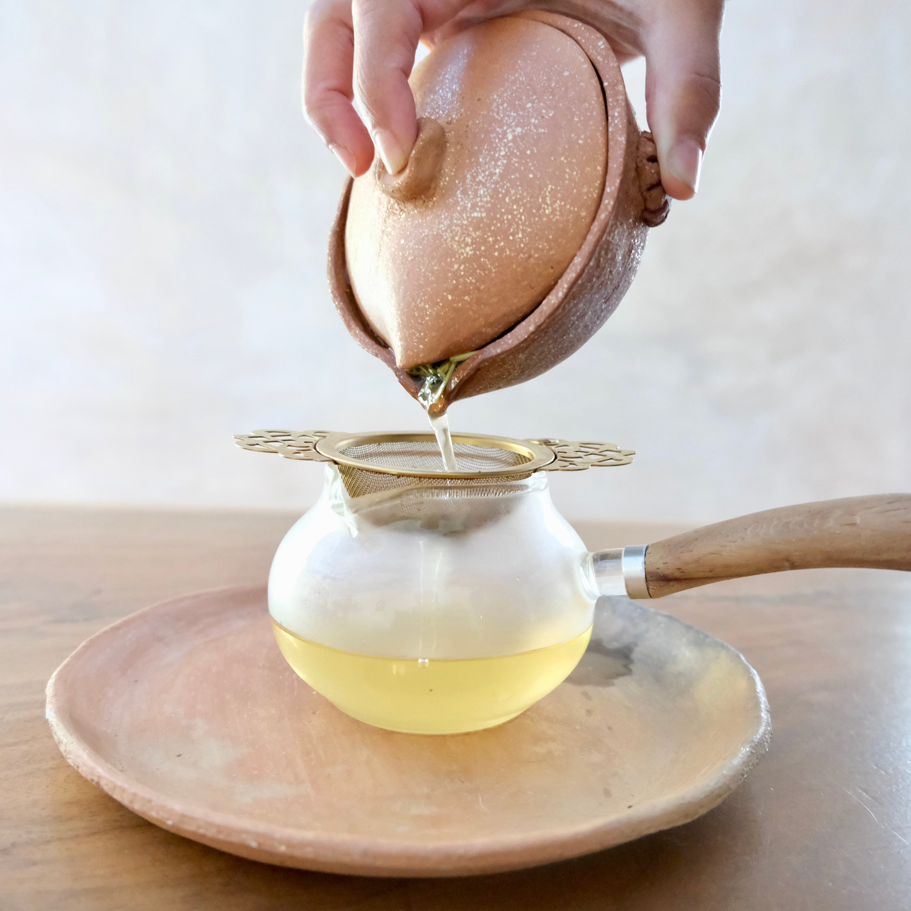 Tea being poured from a ceramic teapot with wooden handle into a glass container on a light background.