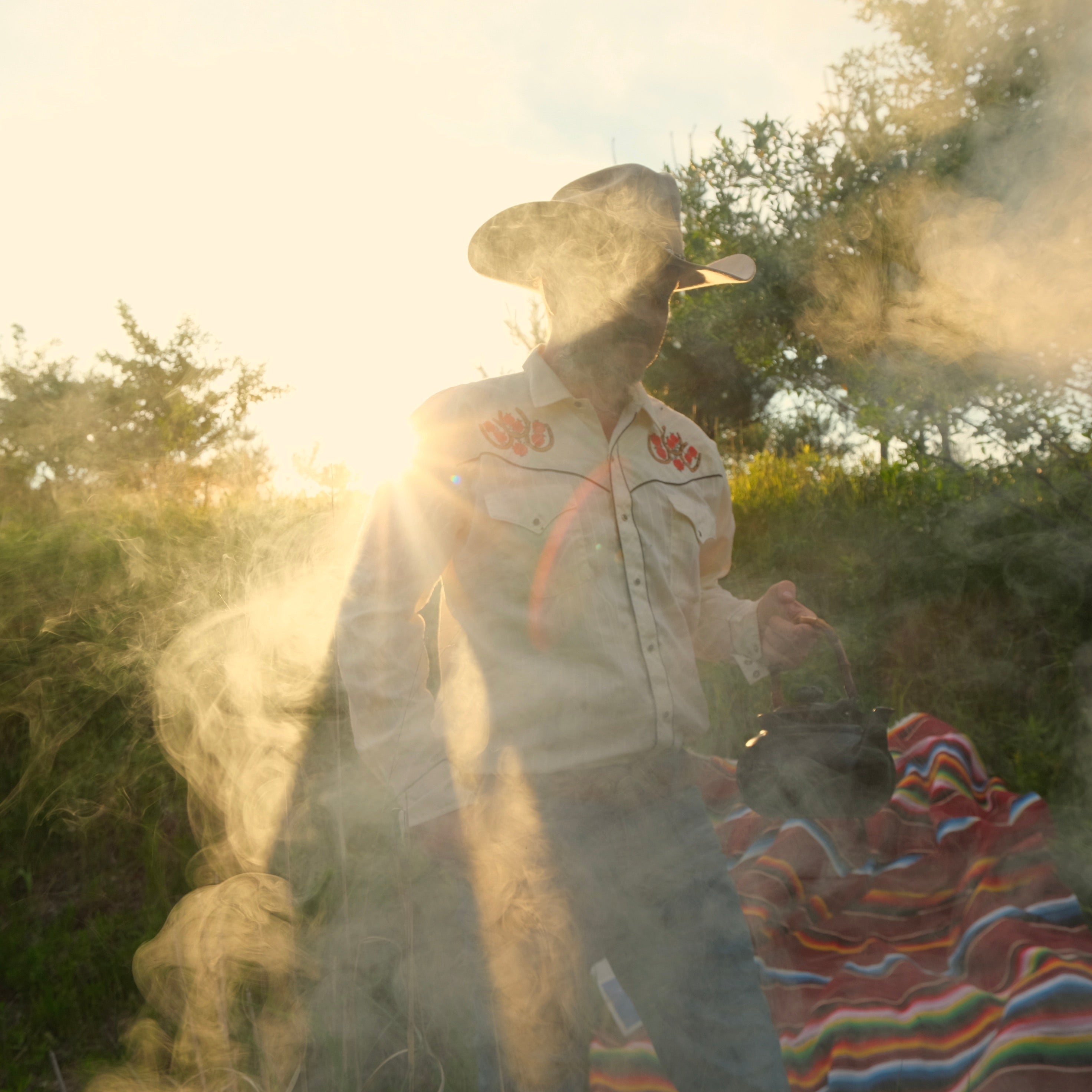 Person in a cowboy hat and patterned shirt standing outdoors with smoke and trees in the background