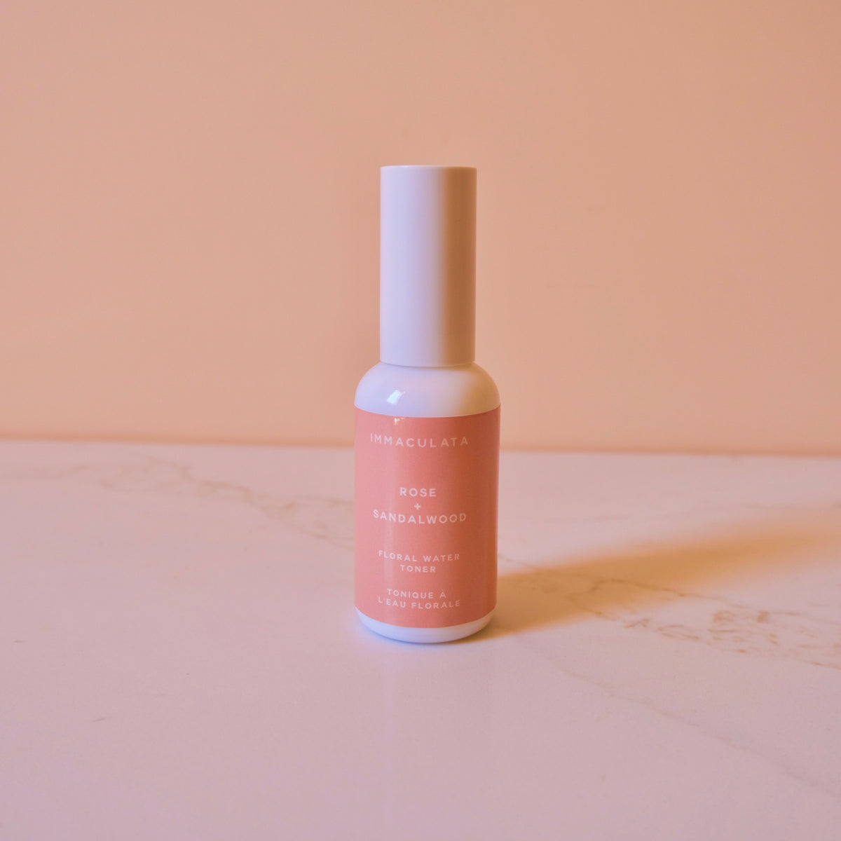 A small bottle of Immaculata Rose Sandalwood Facial Wash & Toner with a white cap and a pink label, standing on a light marble surface against a solid peach background.