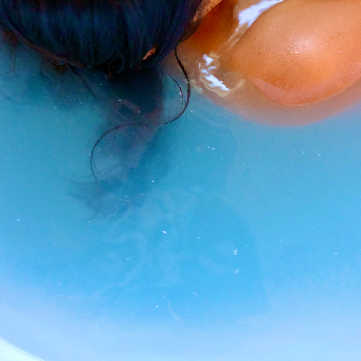 A close-up shot of a person's dark hair and bare shoulder submerged in bright blue water, likely in a bath or pool.