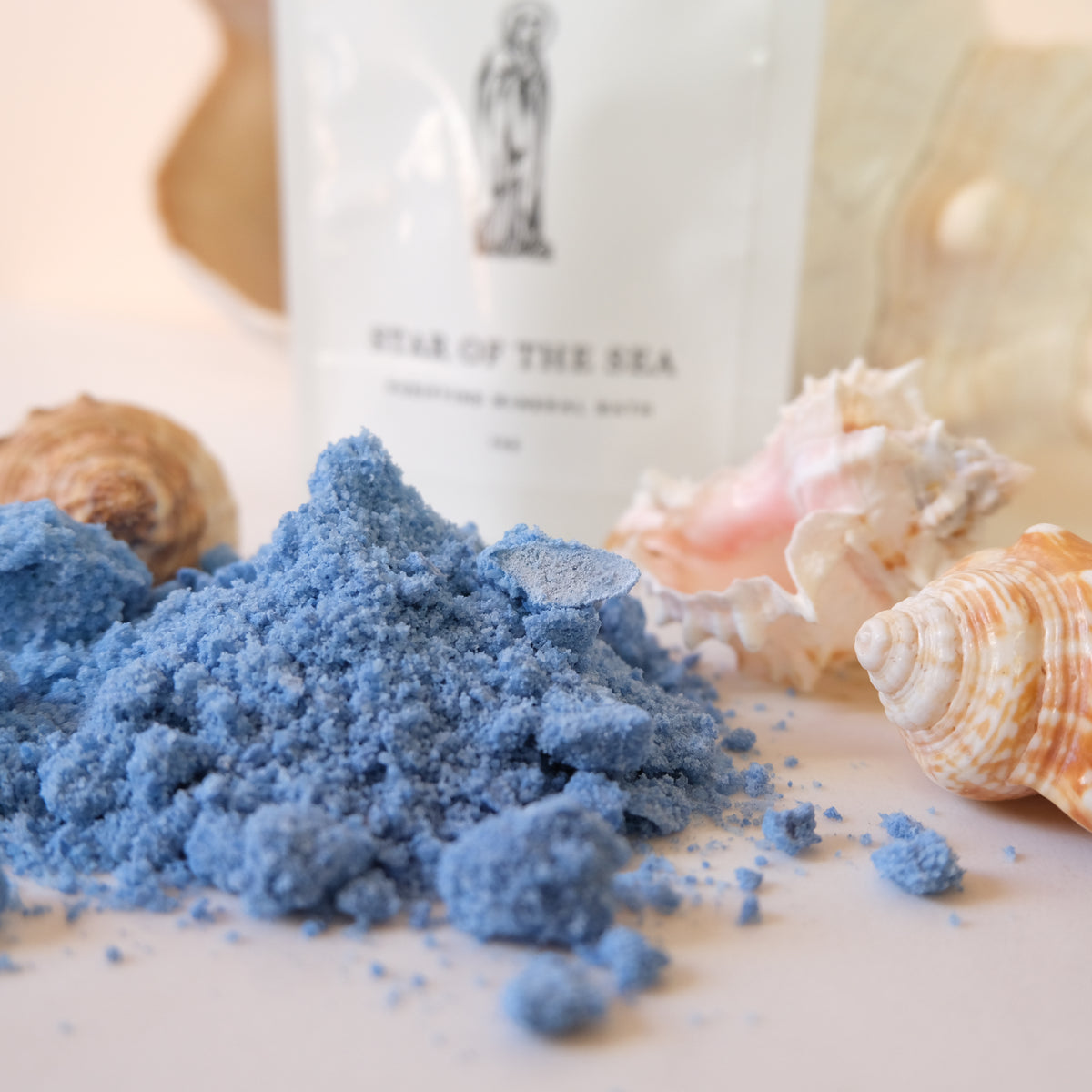 A close-up of bright blue powder, likely sea salt or bath salts, spilled in a small pile on a light surface, with seashells around it and a white pouch labeled 'STAR OF THE SEA' in the background.