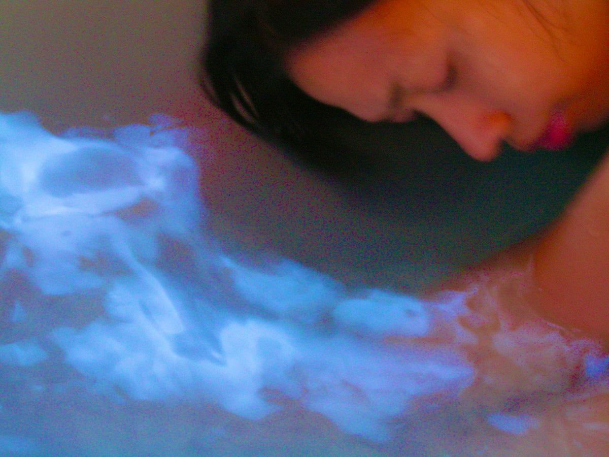 A close-up lifestyle shot of a woman with dark hair resting her head on the edge of a bathtub filled with glowing blue and white bath salts or liquid.