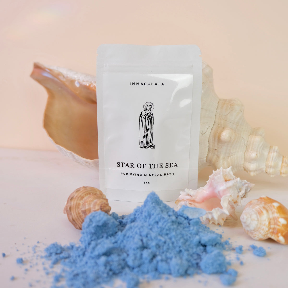 A white stand-up pouch labeled IMMACULATA STAR OF THE SEA PURIFYING MINERAL BATH stands on a light surface with blue bath powder spilled in front and seashells around it against a soft peach background.