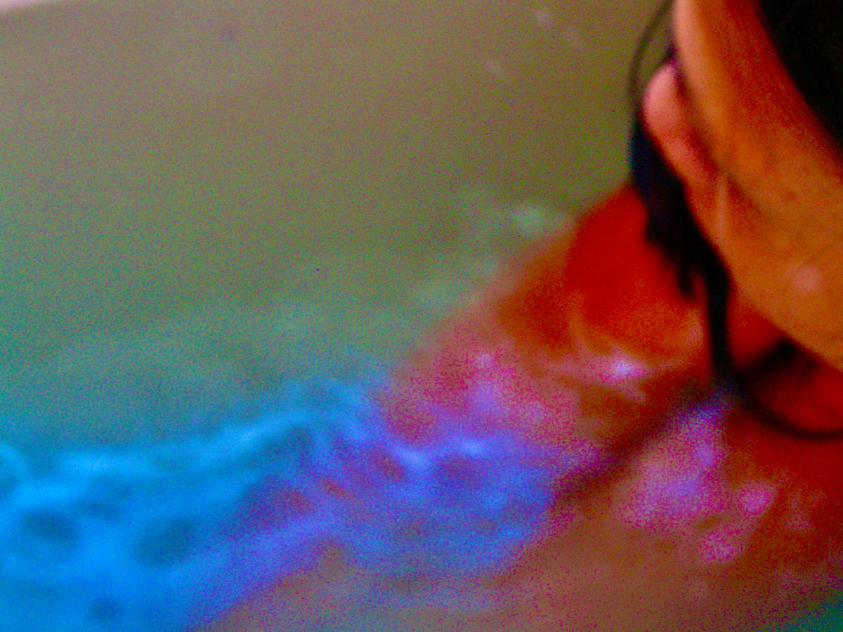 A close-up, grainy shot of a person's face and neck in shallow water, with strong blue, pink, and red color cast effects across the water and skin.