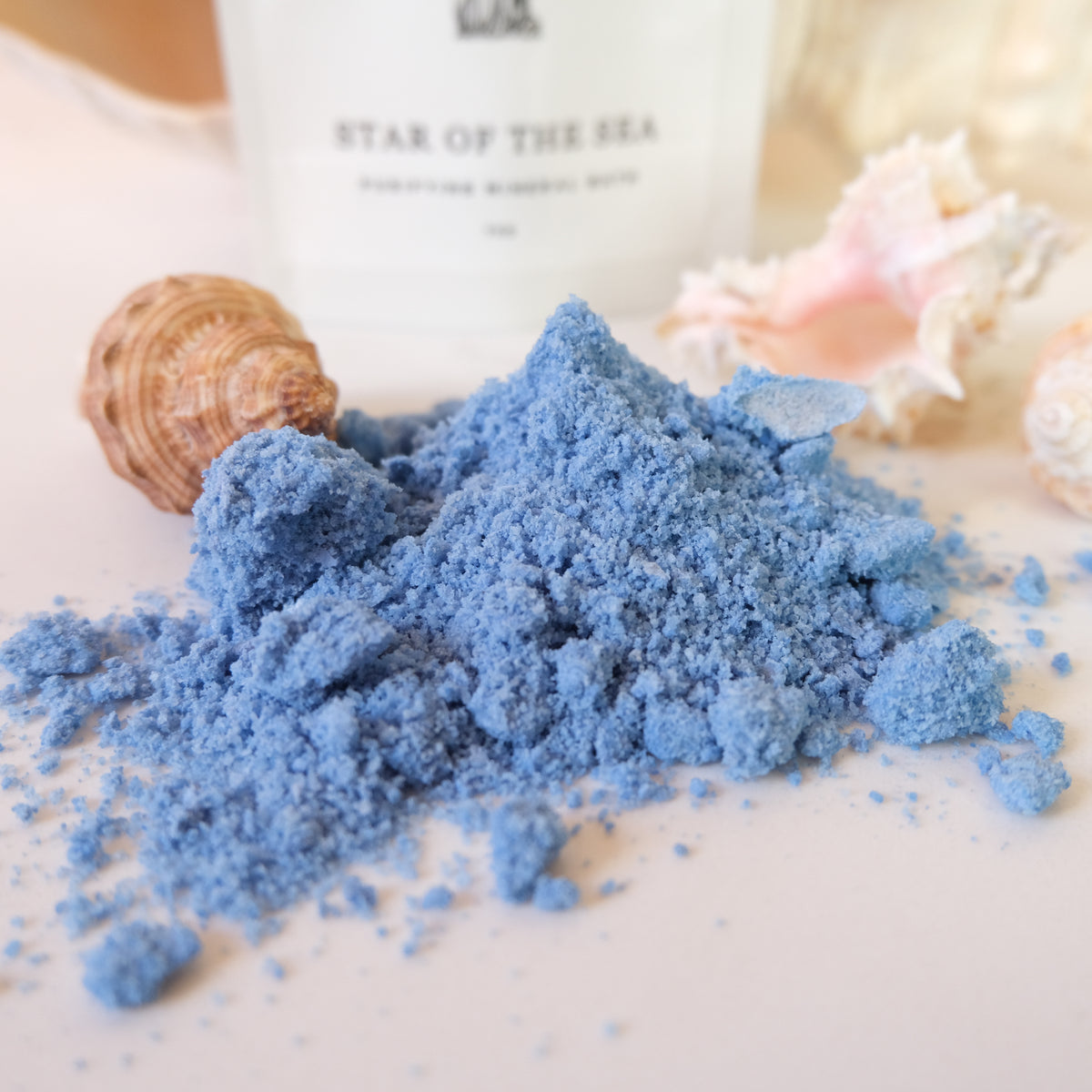 A pile of light blue powder, likely bath salts, displayed on a white surface with seashells and a blurred white bottle in the background. The bottle has the text 'STAR OF THE SEA' and 'PURIFYING MINIMAL BATH' visible.