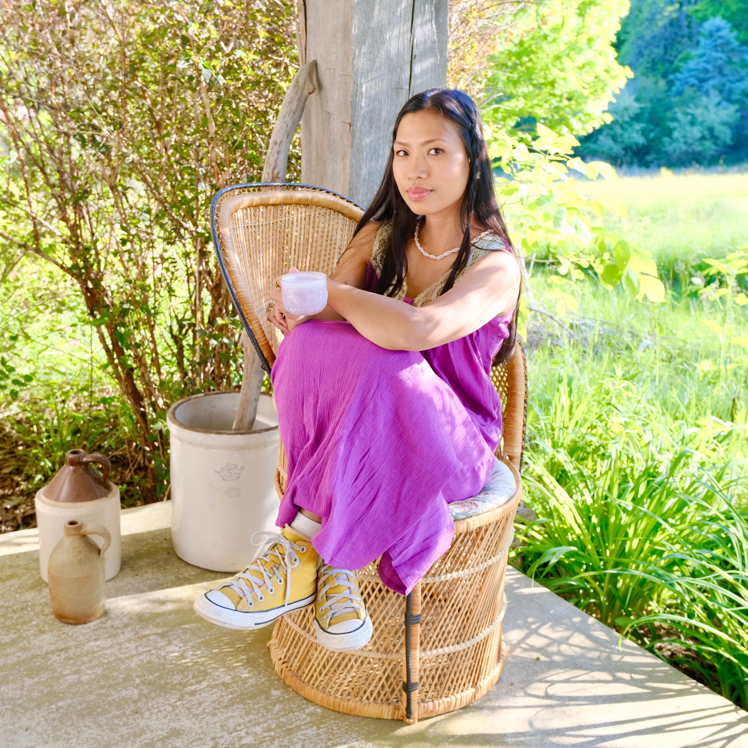 Woman in a purple dress sitting on a wicker chair outdoors with greenery in the background