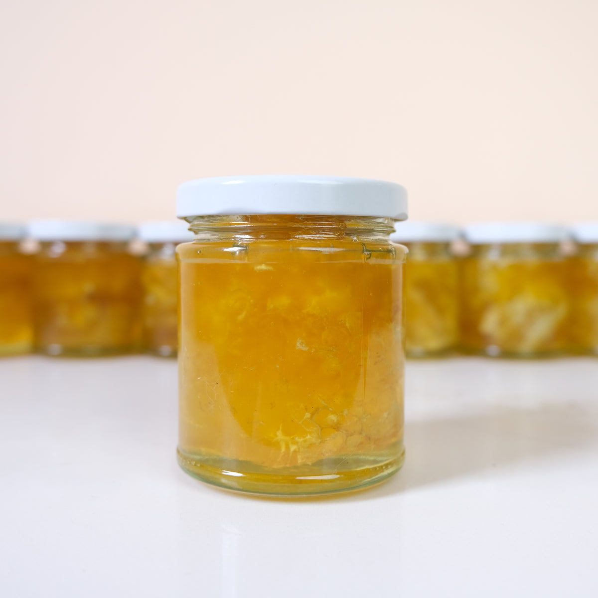 A clear glass jar filled with golden yellow honey, sealed with a white lid, standing on a white surface with several identical jars softly blurred in the background.