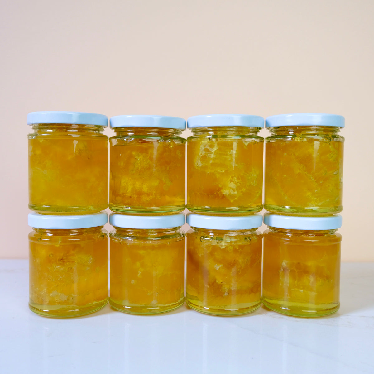 Eight glass jars filled with golden yellow honey, arranged in two rows of four on a white surface against a light beige background.