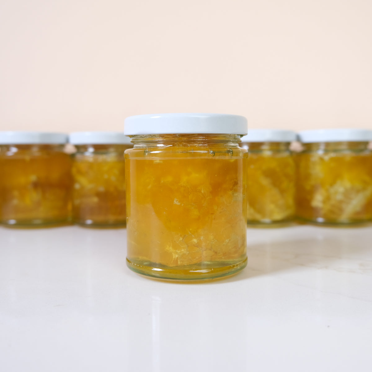 A clear glass jar filled with chunky orange-yellow fruit spread or jam, centered on a white surface with several matching jars blurred in the background against a pale peach wall.