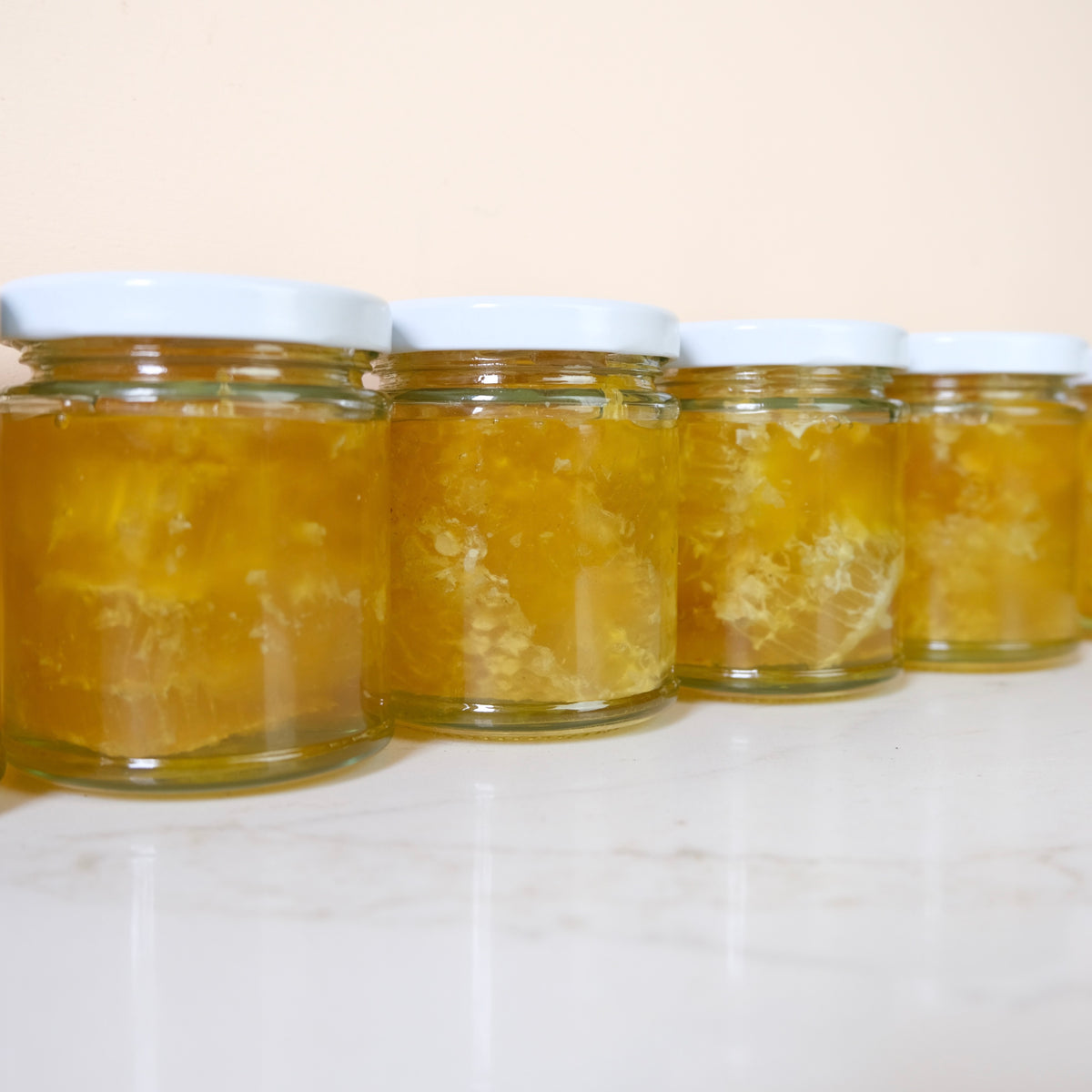 Five small glass jars filled with golden yellow honey, arranged in a row on a white marble surface against a light beige background.