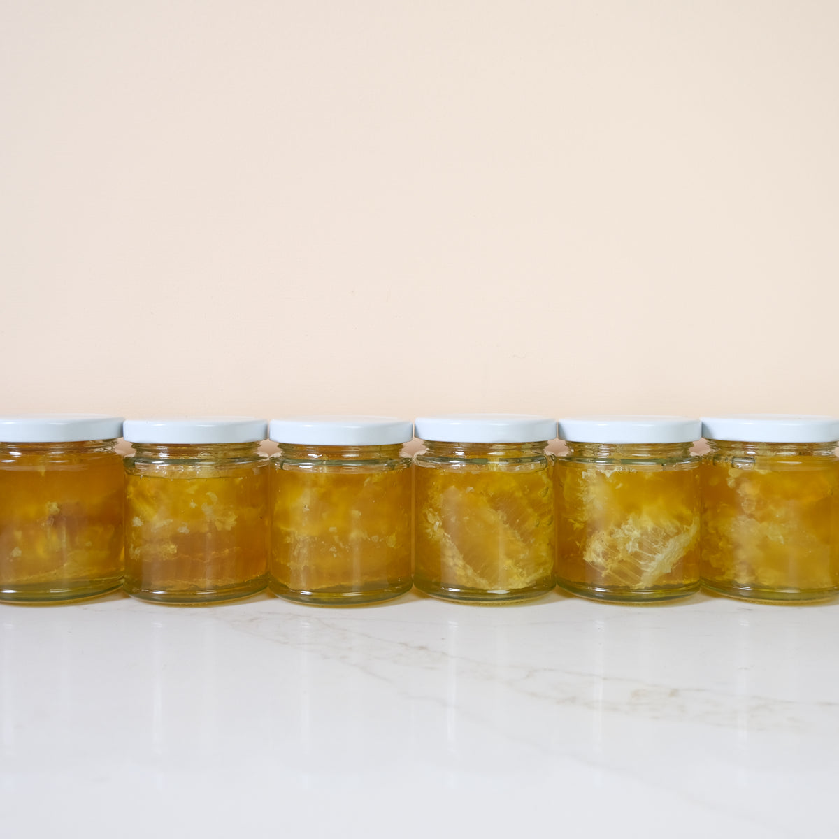 Six glass jars filled with golden honey and visible honeycomb pieces, arranged in a row on a white marble surface against a soft beige background.