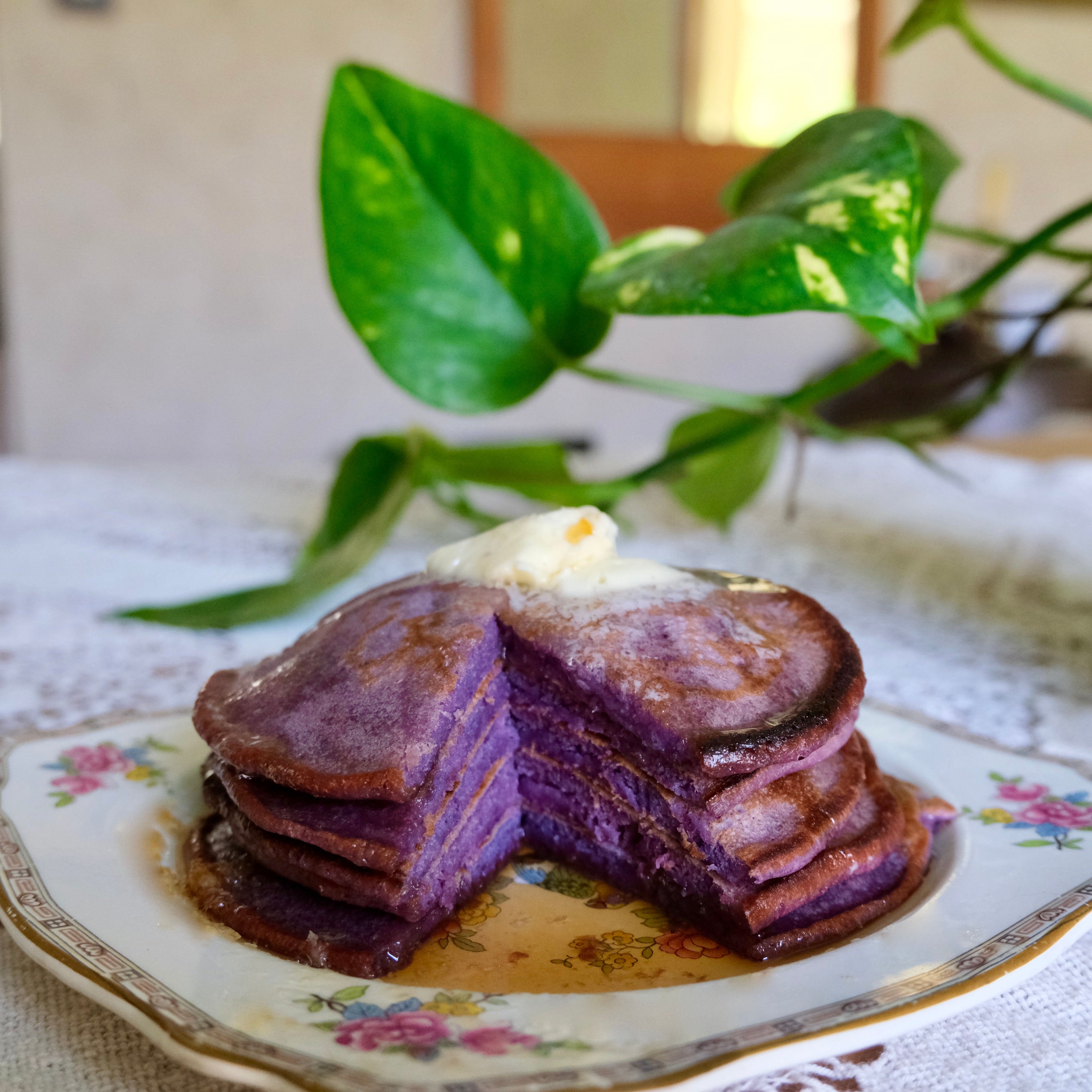 Stack of purple pancakes with a dollop of white cream on a decorative plate, with a plant in the background.
