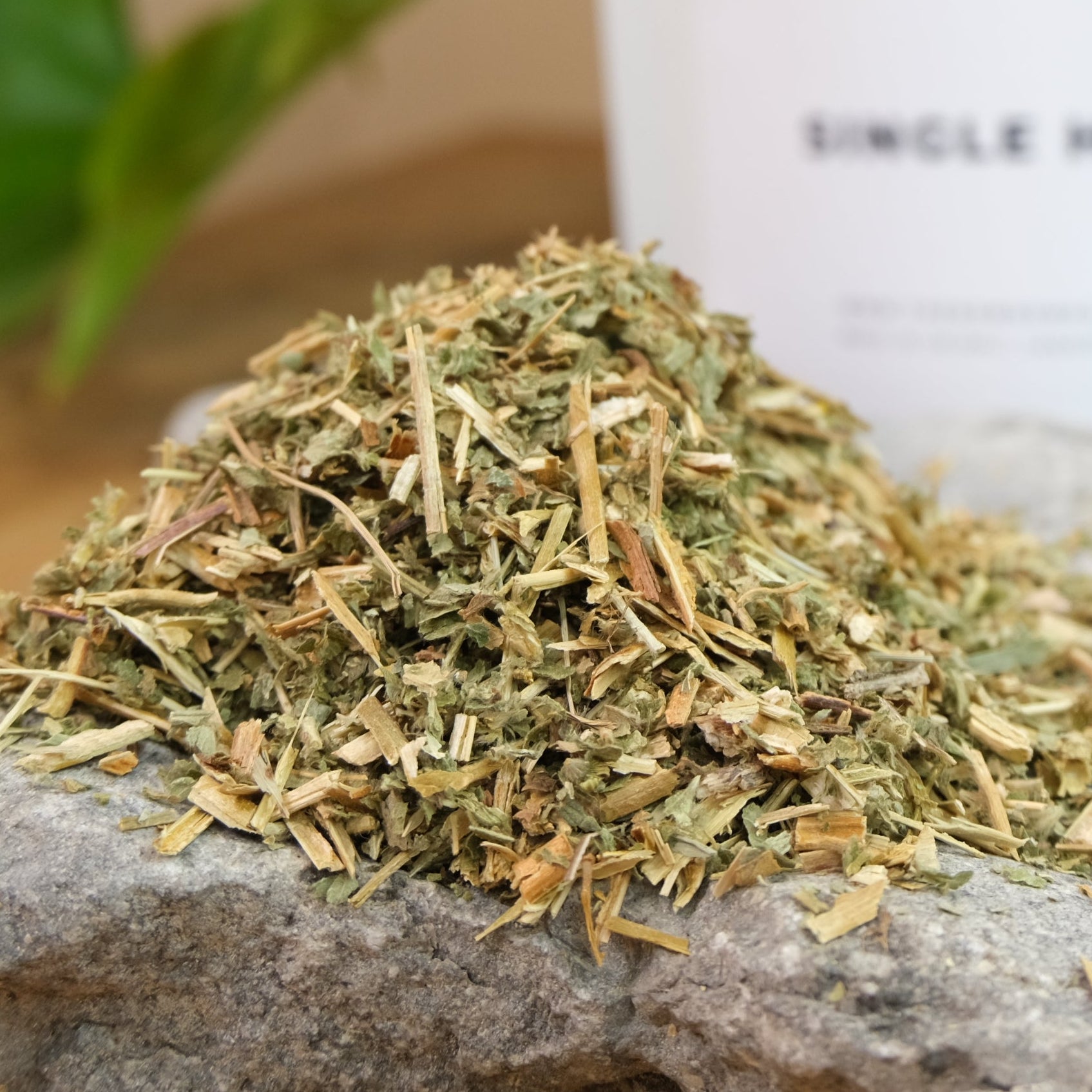 A pile of dried agrimony leaves next to a package labeled 'AGRIMONY' on a rock surface with a plant in the background.