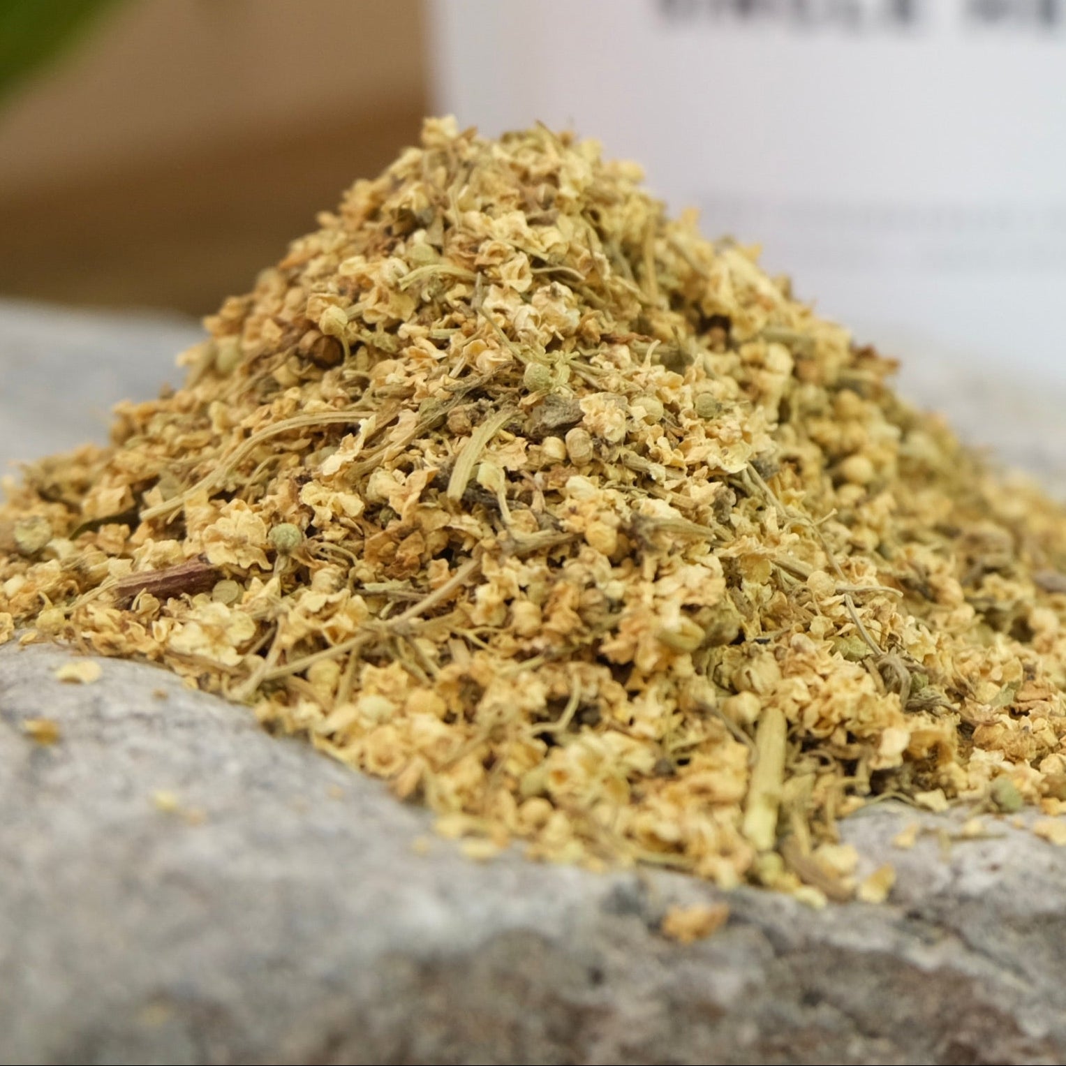 A small pile of dried elderflower tea leaves on a rock, with a white label in the background that reads 'ELDER FLOWER' and 'single herb'.