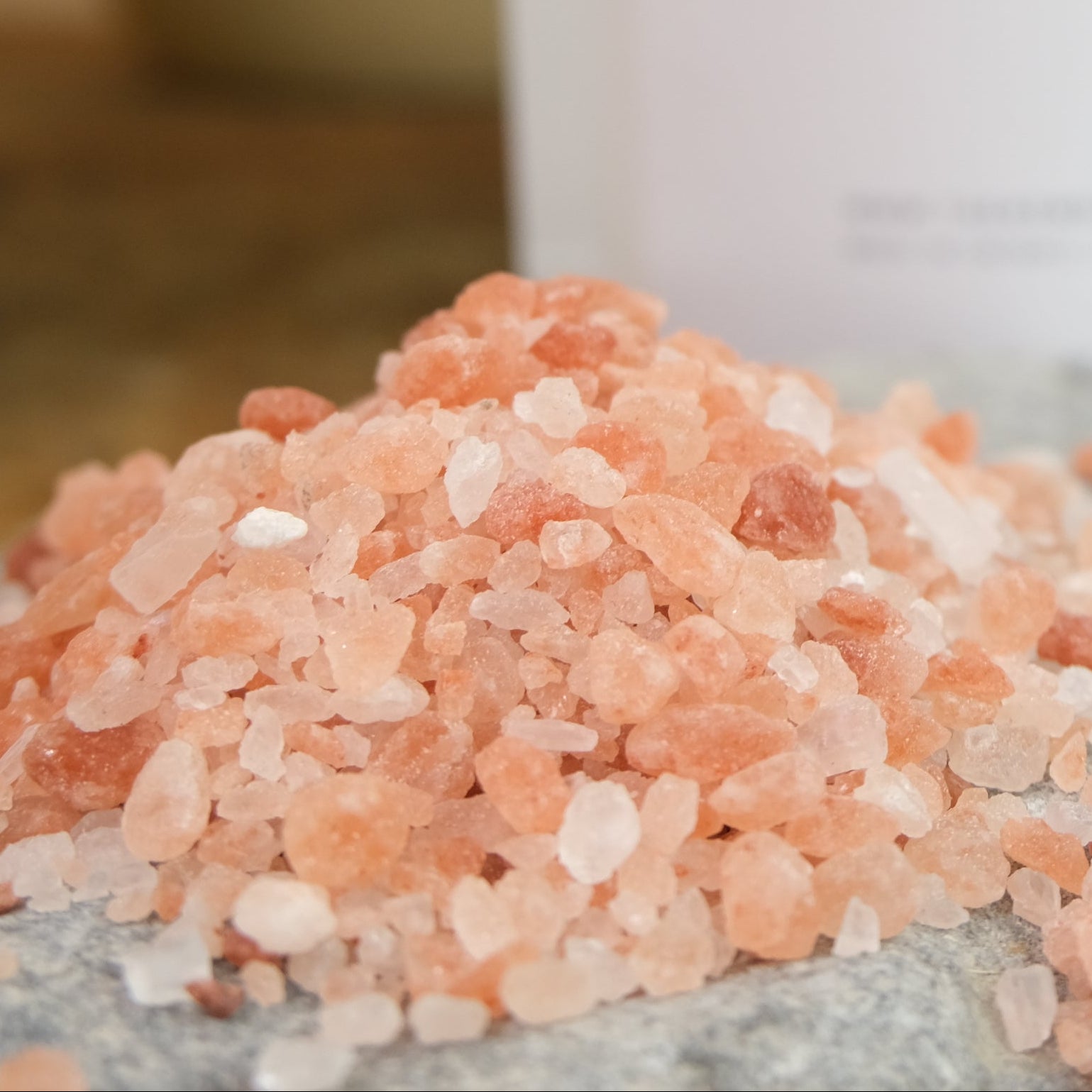 A small pile of pink Himalayan salt crystals in front of a package labeled 'HIMALAYAN PINK SALT'. The salt is on a rock surface with a plant in the background.